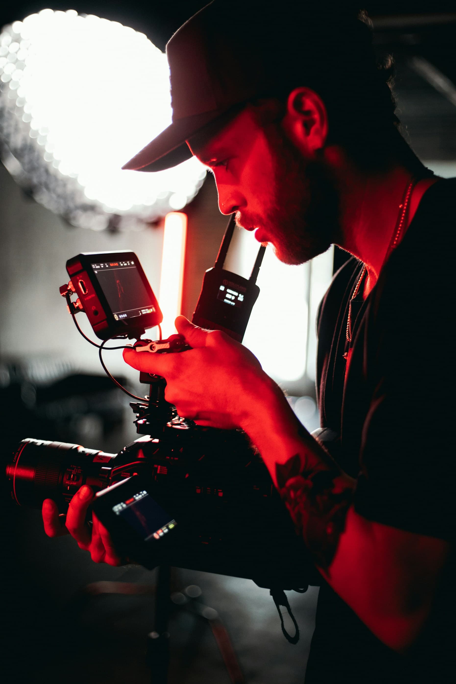 A man in a cap operates a professional camera with multiple screens and attachments, illuminated by red lighting in a dim setting.