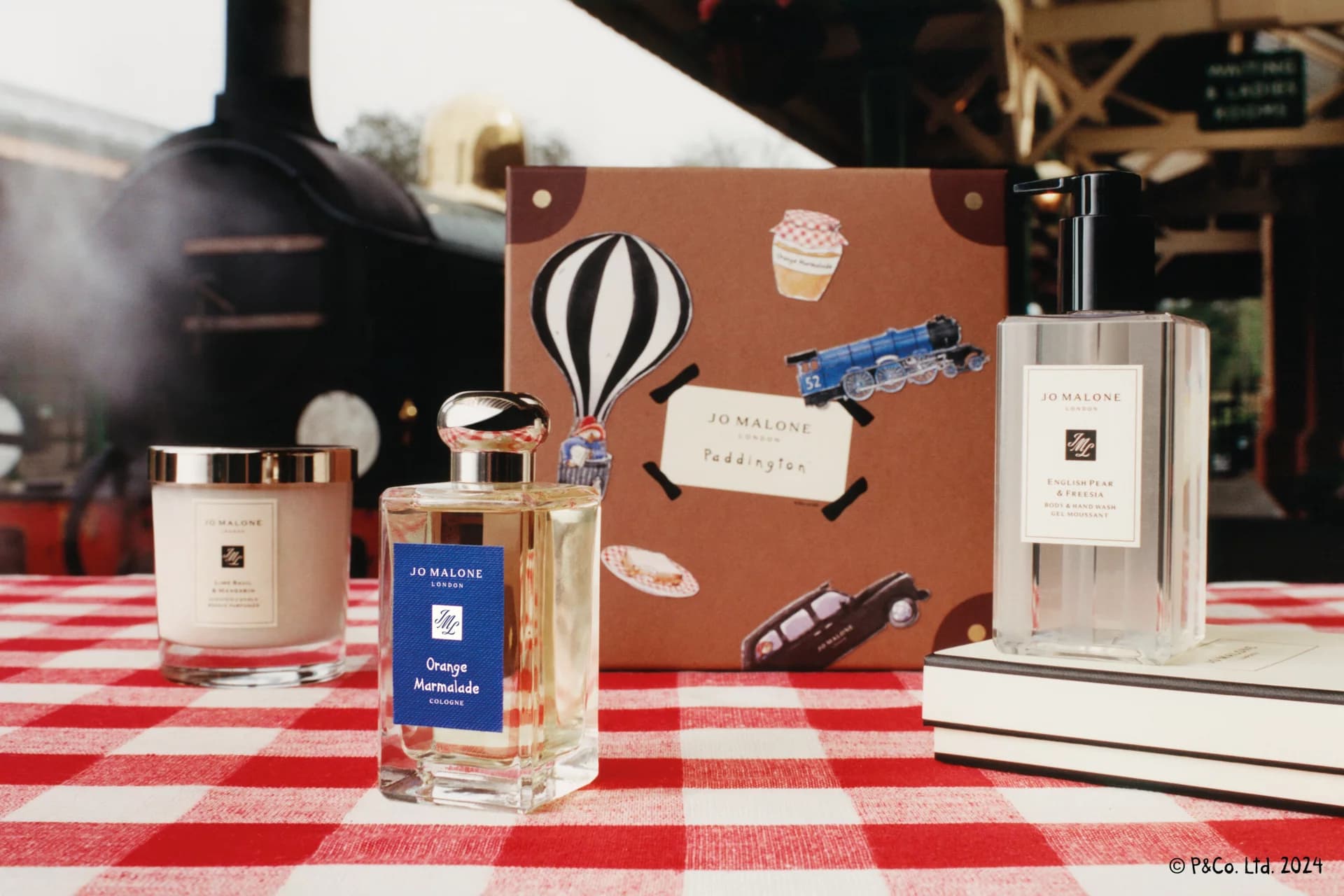 Jo Malone products on a red checkered tablecloth, with a Paddington-themed box featuring a train and hot air balloon design in the background.