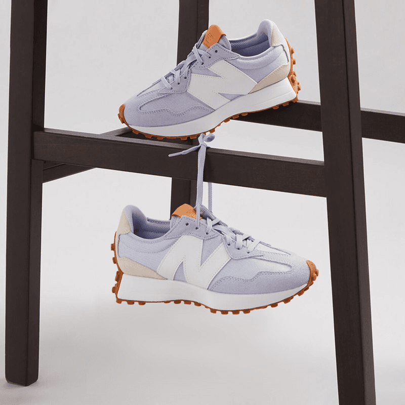 Lilac sneakers with white accents and brown soles are tied together by the laces and hung over a dark wooden chair rung.