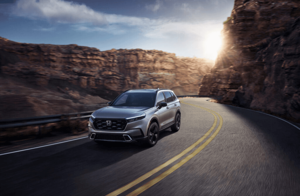 Silver SUV driving on a winding road through a canyon at sunset, with towering rock formations and a clear sky in the background.